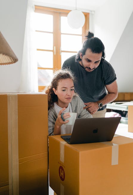 A male worker with short brown hair and a beard, dressed in a blue work uniform, is sitting on the edge of an open white moving van located on a paved driveway. He is holding a clipboard and a pen, ap