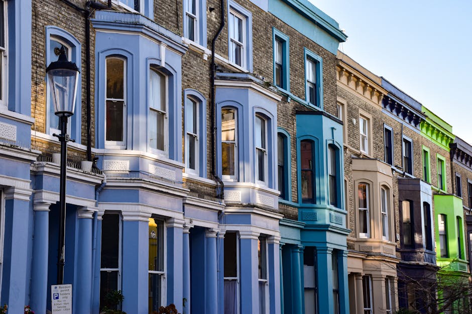 Photograph of a row of multicoloured Victorian terraced houses with bay windows, decorative molding, and brick facades, located on a residential street during daylight. In the foreground, a black traditional street lamp stands next to a parking sign on the sidewalk. The image captures the exterior architecture of the buildings, emphasizing their varied colors, including shades of light blue, teal, beige, black, and bright green, with some houses featuring window frames and detailing in contrasting colors. The windows are mostly double-glazed, with some open or closed, and a few buildings have small front gardens or planters near the entrance. The street is clear, with no visible pedestrians or vehicles, and the overall scene reflects an urban residential environment suitable for house removals or moving services, such as those offered by Removal Companies Ealing, engaged in home relocation or furniture transport in the Ealing borough.
