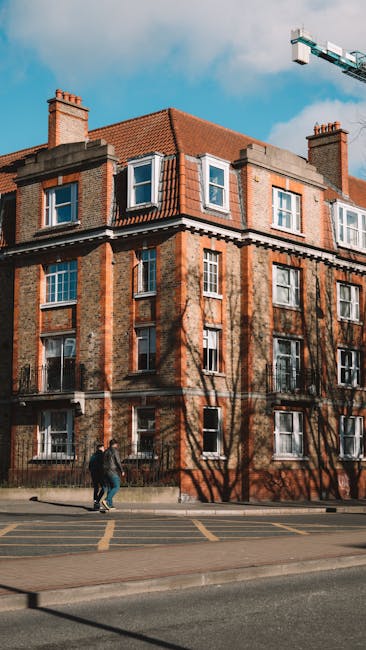 A multi-story residential building constructed of red and brown bricks, with a red-tiled sloped roof featuring dormer windows and chimney stacks. The building has several white-framed sash windows, some open, and black wrought-iron balconies on the upper floors. In front, two people are walking on the pavement near the building, with shadows of leafless trees cast on the facade. A large construction crane is visible in the clear blue sky above. The scene is set in an urban environment, with a street that has visible parking markings, suggesting a location conducive to home relocations and furniture transport. The image reflects a typical town centre setting suitable for house removals and moving services offered by Removal Companies Ealing as part of their comprehensive relocation support.