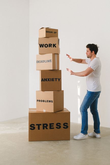 A young man wearing a white t-shirt and blue jeans is standing indoors next to a vertical stack of six cardboard boxes arranged from largest at the bottom to smallest at the top. Each box has a word w