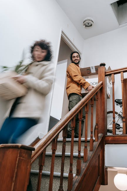 A man with a beard and wearing an orange jacket is standing on a staircase landing inside a home, holding a cardboard box with labels. The staircase has wooden banisters and carpeted steps. A woman with curly hair, dressed in a light-colored coat and blue jeans, is walking past on the staircase, slightly blurred due to motion. In the background, there is a doorway leading to another room, and part of a bicycle is visible further upstairs. The ceiling has a smoke detector, and the walls are painted white. The scene captures the process of home relocation, with furniture and boxes being moved, supported by a professional removal service such as Removal Companies Ealing. The lighting is natural and well-lit, indicating daylight, and the overall environment reflects a typical residential interior during a packing and moving operation.