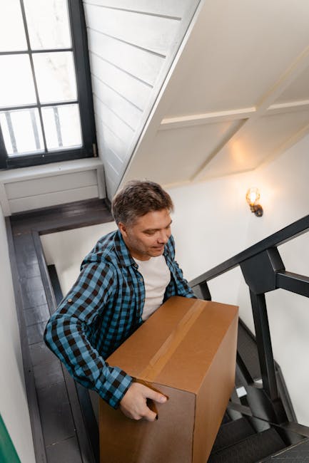 A man with short brown hair wearing a blue checkered shirt is carrying a medium-sized rectangular cardboard box up a staircase inside a home. The staircase has dark wooden steps and black metal railing, and the walls are painted white with decorative molding. A window with black framing is visible at the top of the stairs, allowing natural light to illuminate the area, while an indoor wall sconce provides additional lighting. This scene illustrates the process of home relocation and furniture transport, with the individual preparing to move the box through a narrow staircase. Slightly visible in the background is a door and part of a white panel wall, emphasizing the interior environment. The image reflects professional moving activities carried out by Removal Companies Ealing, focusing on packing and logistical aspects of house removals through staircases.