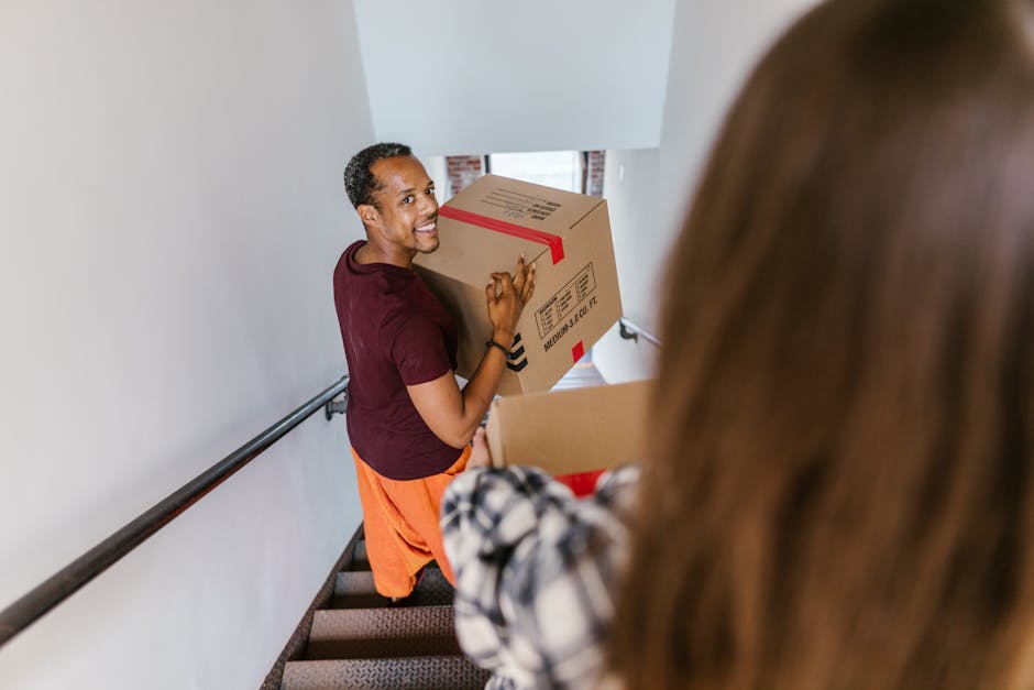 A man with a beard and wearing an orange jacket is standing on a staircase landing inside a home, holding a cardboard box with labels. The staircase has wooden banisters and carpeted steps. A woman with curly hair, dressed in a light-colored coat and blue jeans, is walking past on the staircase, slightly blurred due to motion. In the background, there is a doorway leading to another room, and part of a bicycle is visible further upstairs. The ceiling has a smoke detector, and the walls are painted white. The scene captures the process of home relocation, with furniture and boxes being moved, supported by a professional removal service such as Removal Companies Ealing. The lighting is natural and well-lit, indicating daylight, and the overall environment reflects a typical residential interior during a packing and moving operation.