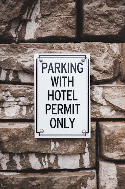 A rectangular white metal sign with black text reading 'PARKING WITH HOTEL PERMIT ONLY' is mounted on a stone wall constructed of large, irregularly shaped brownish-beige stones. The sign is secured with four bolts, one at each corner. In the foreground, part of a home relocation and furniture transport process is visible, with several cardboard boxes wrapped in plastic and fabric packing materials stacked inside an entrance or on a pavement near the building. A hand truck or trolley can be seen partially, used for loading or unloading boxes into or from a van parked nearby. The scene depicts the organized moving process, including packing materials and the logistics of parking permits for a relocation service provided by the local removal company, Removal Companies Ealing. Bright natural light illuminates the scene, highlighting the textures of the stone wall and the packing supplies, emphasizing the context of a professional home or office move in an urban setting.