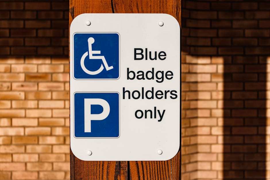 A white rectangular parking permit sign mounted on a wooden post in front of a brick wall shows a blue wheelchair accessibility symbol at the top and a blue parking symbol at the bottom. The sign has black text that reads 'Blue badge holders only' aligned to the right of the symbols. The image is taken outdoors, with clear daylight illuminating the sign and surroundings. The setting suggests an urban environment, possibly near a residential or commercial property, where parking for disabled individuals is designated. This type of signage is relevant for home relocation or furniture transport services, such as those provided by Removal Companies Ealing, to ensure proper parking arrangements during a house removal or moving process beyond the immediate loading area.