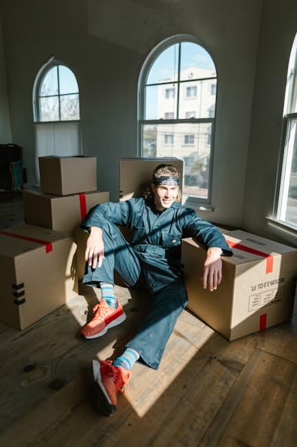 A young man with long hair, wearing a blue jumpsuit, orange sneakers, and striped socks, sits on the wooden floor in a bright room with large arched windows. Behind him are several large cardboard moving boxes with red and black tape, some stacked and some placed on the floor, indicating an active home relocation process. The room receives natural daylight through the windows, which offer a view of neighboring buildings. The scene captures the preparation stage of packing and moving, with the individual taking a break during furniture transport or packing activities associated with a same-day removal service. This setting is typical of house removals and moving logistics handled by Removal Companies Ealing.