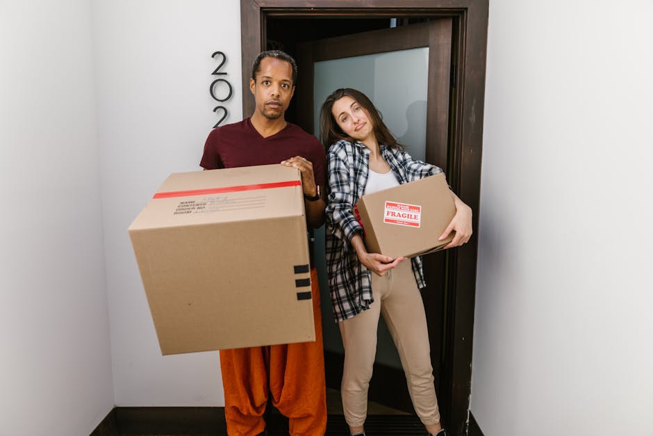 Two individuals, a man and a woman, stand inside an apartment doorway, each holding a cardboard box prepared for home relocation. The man, positioned on the left, has dark hair and wears a maroon t-shirt and orange trousers, holding a large box in front of him. The woman, on the right, has long brown hair and is dressed in a plaid shirt over a white top with beige trousers, holding a smaller box marked with a red 'Fragile' sticker. The doorway features a dark wooden frame with a frosted glass panel, and the interior walls are white, suggesting a clean, modern environment. The scene captures the loading process typical of professional removals, with boxes ready for transport by moving service providers such as Removal Companies Ealing. Visible floor space and part of a corridor hint at the initial stages of a home or apartment move, emphasizing packing and furniture transport tasks involved in same day removals in W5, with a focus on careful handling of fragile items and efficient moving logistics.